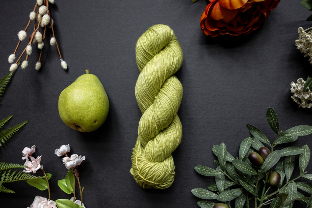 A bright green skein of sock weight yarn lies on a black surface. It's surrounded by flowers, branches, an orange rose, and a pear.