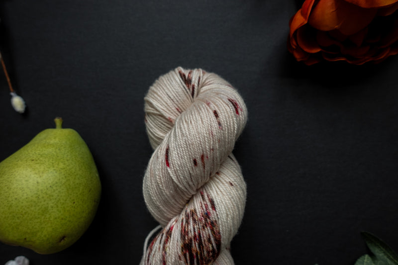 Seen close up, a naturally dyed skein of white yarn with burgundy speckles lays on a black surface next to an orange-red flower and a pear.