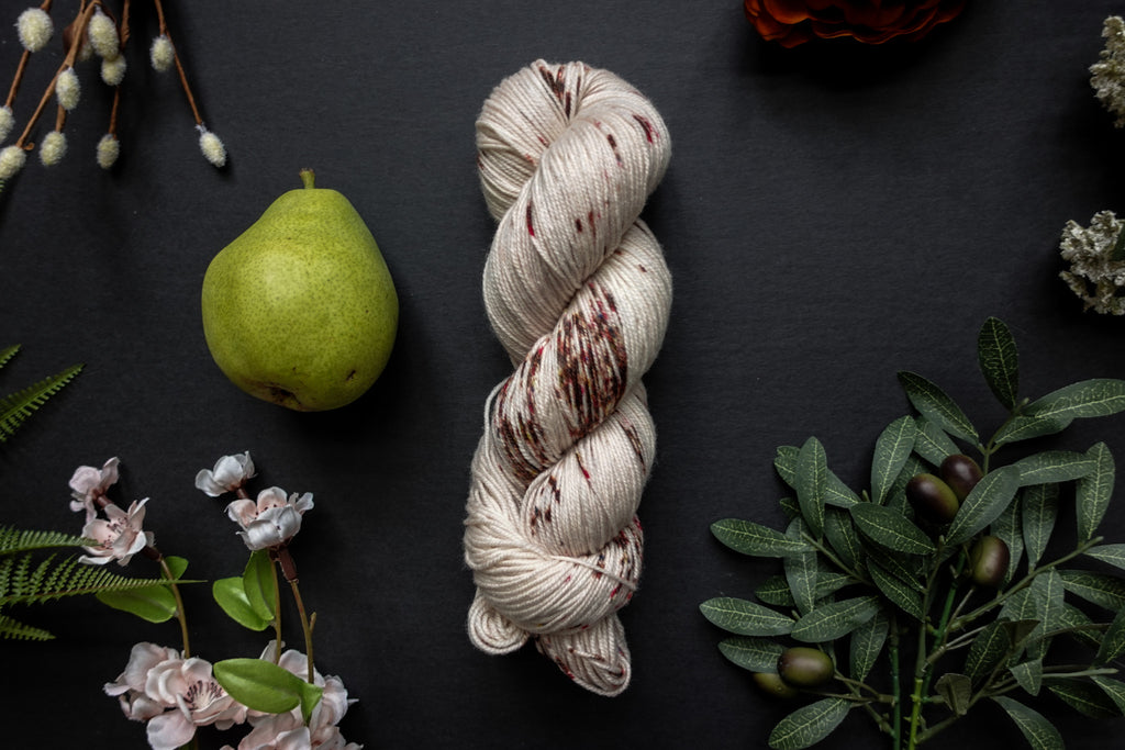 A skein of white sock weight yarn with burgundy speckles lies on a black surface. It's surrounded by flowers, branches, an orange rose, and a pear.