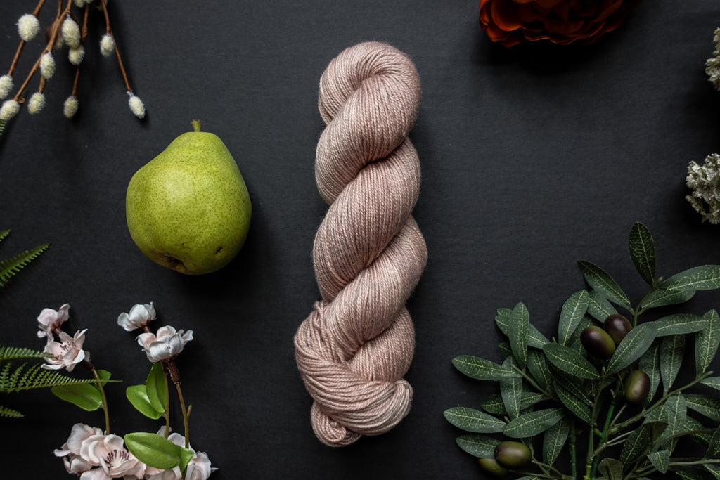 A skein of light beige sock weight yarn lies on a black surface. It's surrounded by flowers, branches, an orange rose, and a pear.