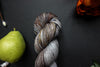 Seen close up, a variegated skein of brown, grey, and white naturally dyed yarn lays on a black surface next to an orange-red flower and a pear.