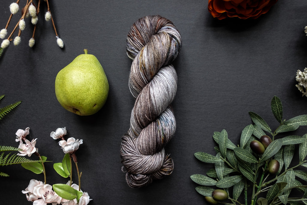 A variegated skein of brown, white, and beige sock weight yarn lies on a black surface. It's surrounded by flowers, branches, an orange rose, and a pear.