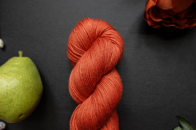 Seen close up, a naturally dyed skein of orange-red yarn lays on a black surface next to a matching flower and a pear.