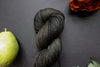 Seen close up, a muddy green, almost black, skein of hand dyed yarn lays on a black surface. It's surrounded by an orange-red flower and a pear.