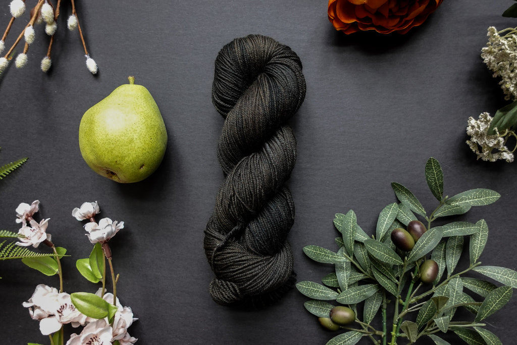 A dark, muddy green, almost black, skein of sock weight yarn lies on a black surface. It's surrounded by flowers, branches, an orange rose, and a pear.