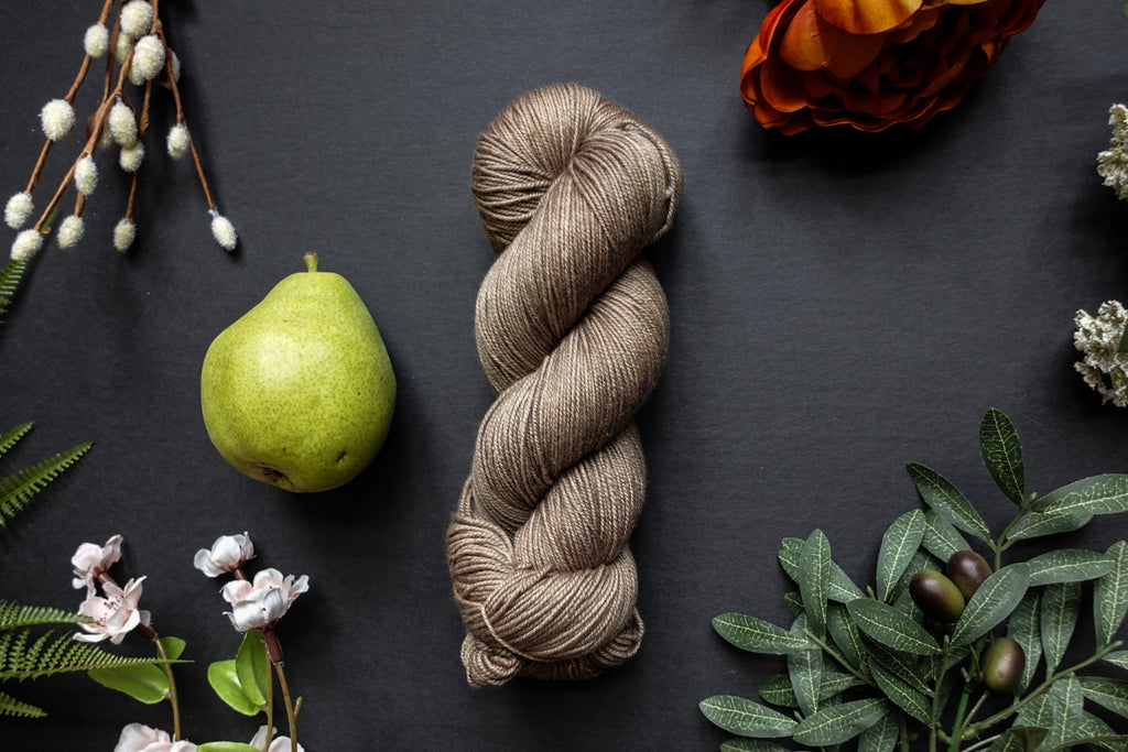 A beige brown skein of sock weight yarn lies on a black surface. It's surrounded by flowers, branches, an orange rose, and a pear.