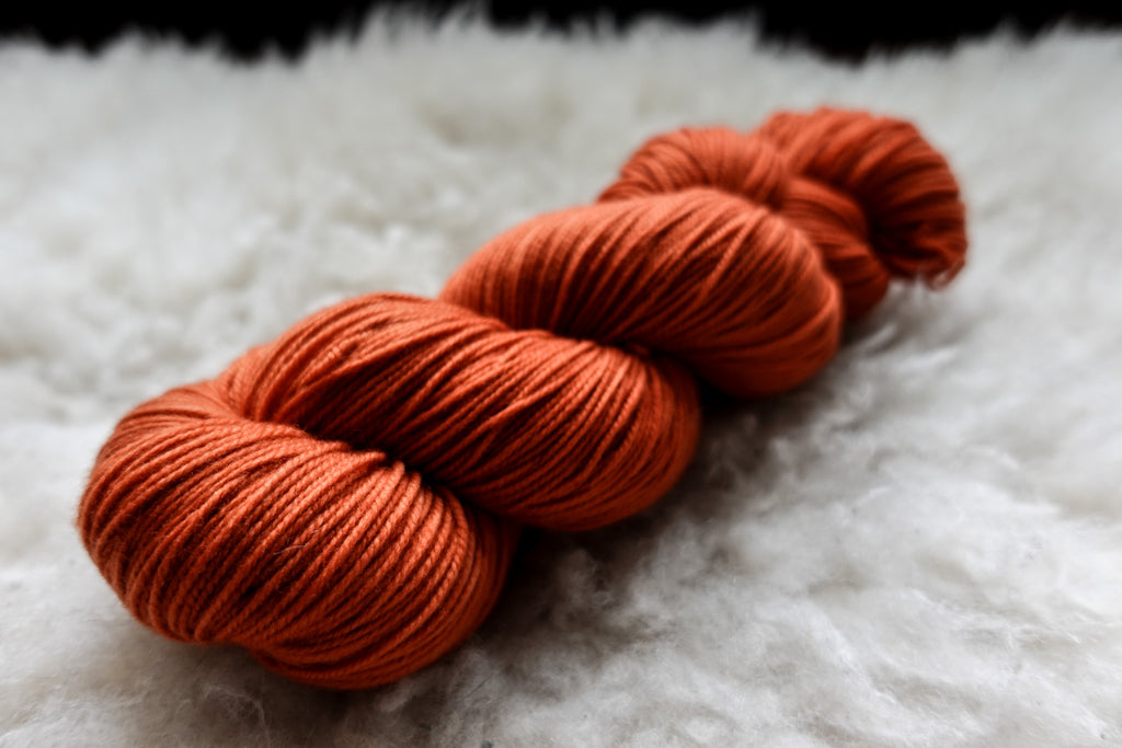 A natural skein of yarn has been hand dyed orange-red. It lays on a sheepskin and is seen from the side.
