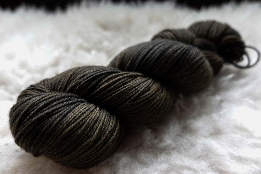 A dark, almost black, skein of natural yarn has been hand dyed. It lays on a sheepskin and is seen from the side.