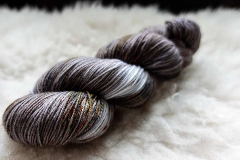 A natural skein of yarn, hand dyed grey, brown, and white, lays on a sheepskin and is seen from the side.