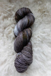 Seen from above, a variegated skein of grey, brown, and white hand dyed yarn lays on a sheepskin.