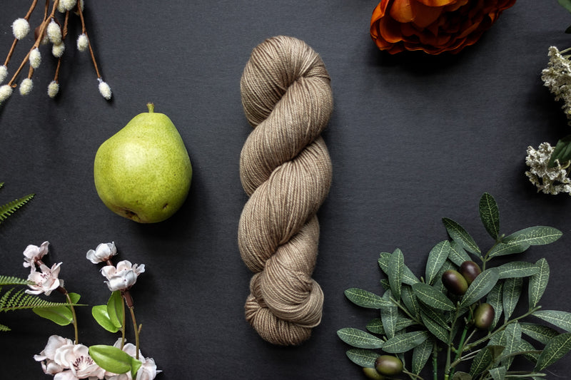 A beige brown skein of sock weight yarn lies on a black surface. It's surrounded by flowers, branches, an orange rose, and a pear.