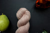 Seen close up, a light pink-beige skein of hand dyed yarn lays on a black surface next to and orange-red flower and a pear.