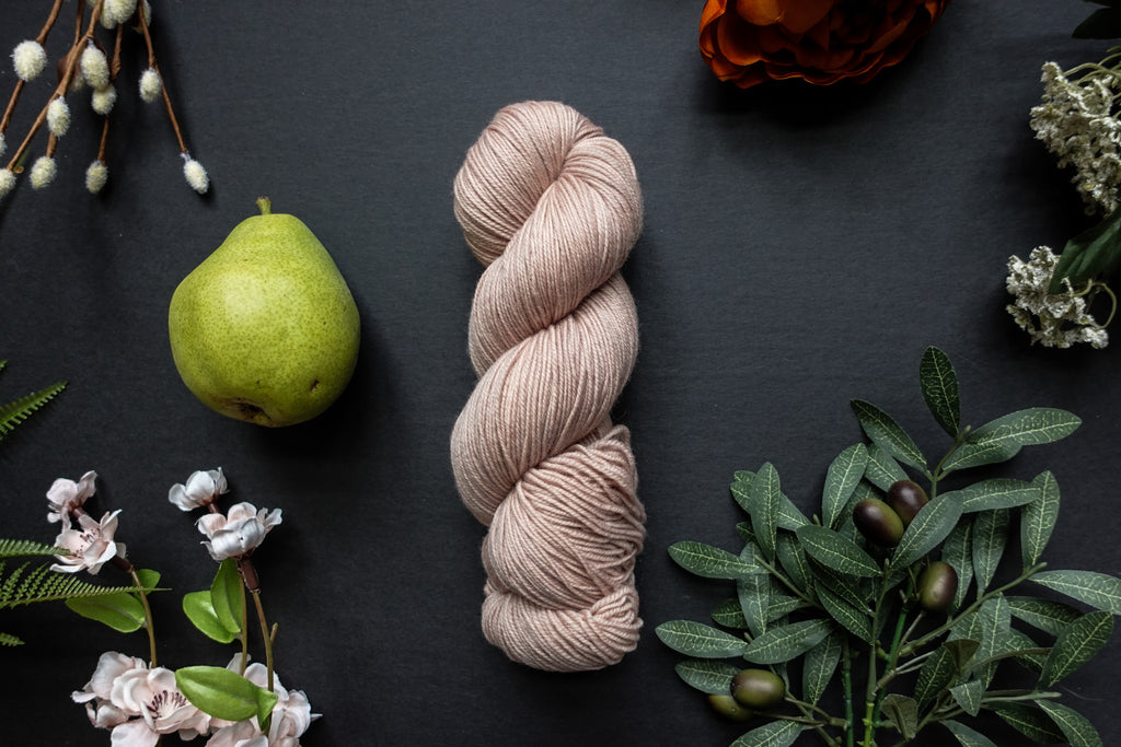 A skein of light beige sock weight yarn lies on a black surface. It's surrounded by flowers, branches, an orange rose, and a pear.