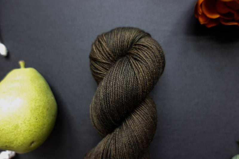 Seen close up, a muddy green, almost black skein of hand dyed yarn lays on a black surface next to an orange-red flower and a pear.