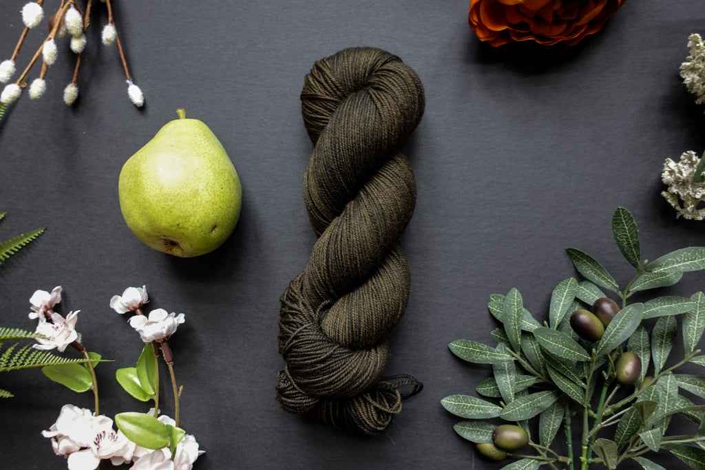 A dark, muddy green skein of sock weight yarn lies on a black surface. It's surrounded by flowers, branches, a poppy, and a pear.