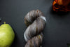 Seen close up, a grey, brown, and white naturally dyed skein of yarn lays on a black surface next to an orange-red flower and a pear.