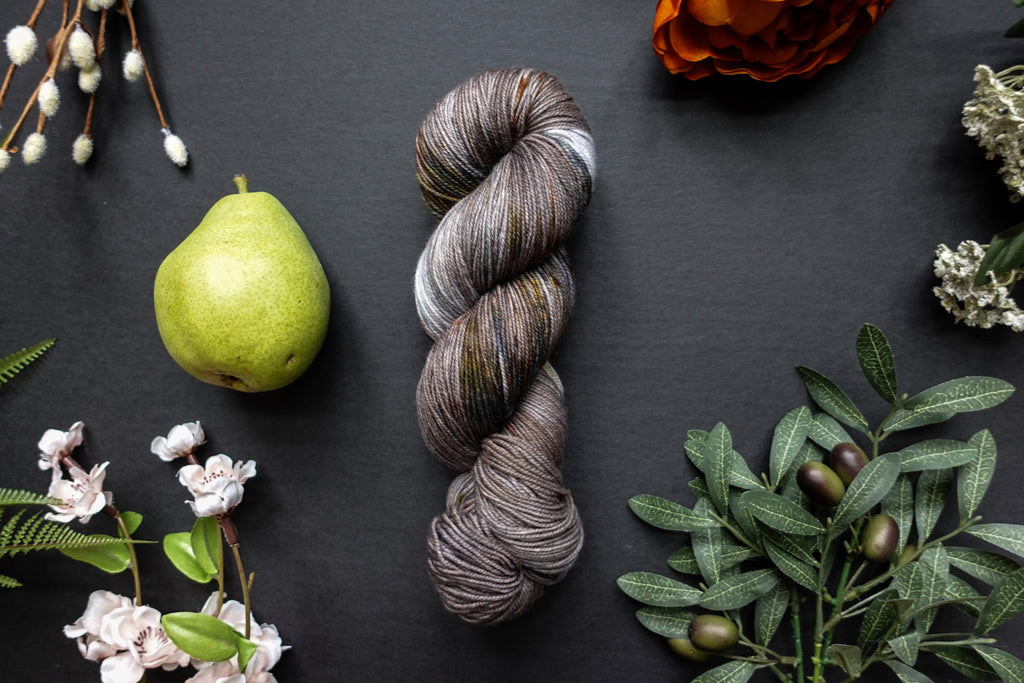 A variegated skein of brown, white, and beige sock weight yarn lies on a black surface. It's surrounded by flowers, branches, an orange rose, and a pear.