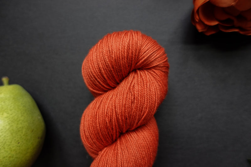 Seen close up, an orange-red skein of hand dyed yarn lays on a balck surface next to a matching flower and a pear.