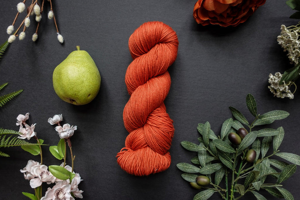 A orange-red skein of sock weight yarn lies on a black surface. It's surrounded by flowers, branches, an orange rose, and a pear.