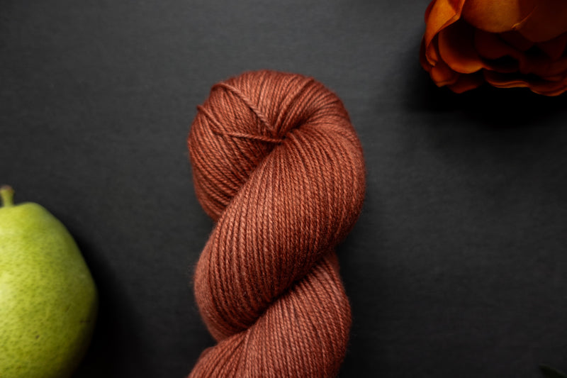 Seen close up, a brick red skein of hand dyed yarn lays on a black surface, next to an orange-red flower and a pear.