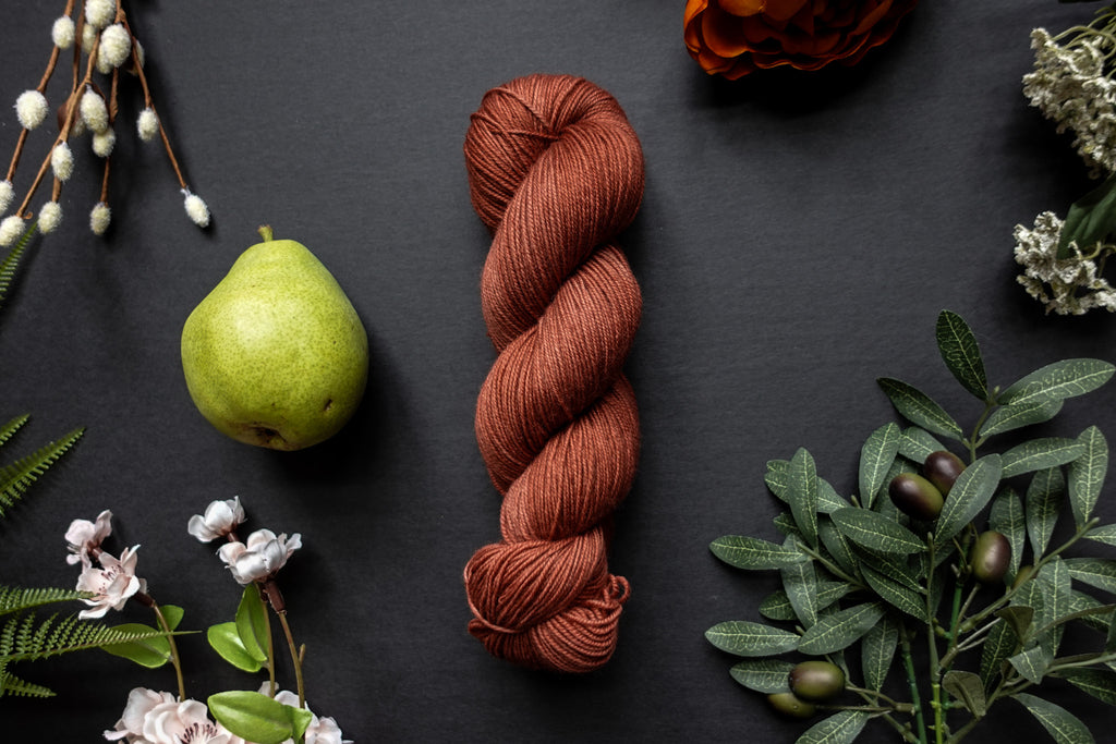 A brick red skein of sock weight yarn lies on a black surface. It's surrounded by flowers, branches, an orange rose, and a pear.