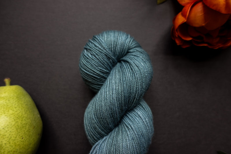 Seen close up, a indigo blue skein of hand dyed yarn lays on a black surface next to an orange-red flower and a pear.
