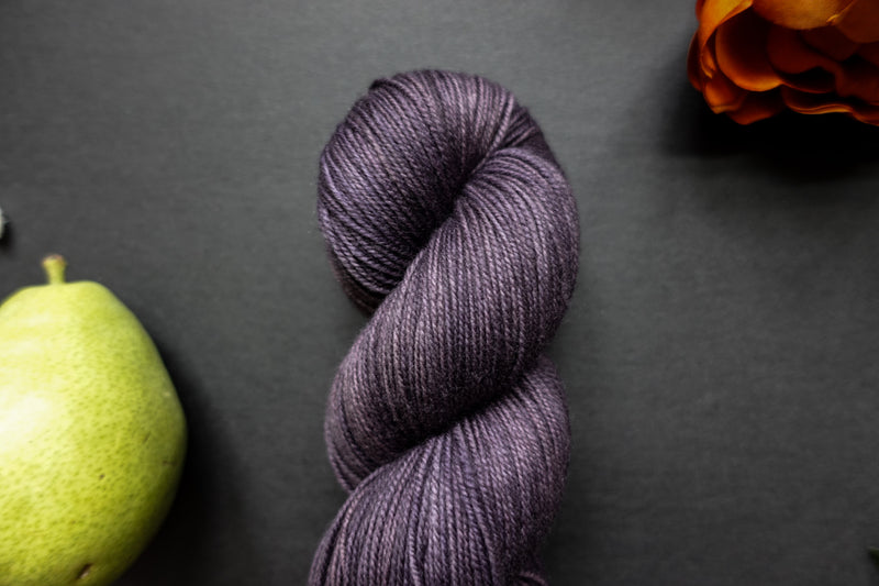 Seen close up, a deep purple skein of naturally dyed yarn lays on a black surface next to an orange-red flower and pear.