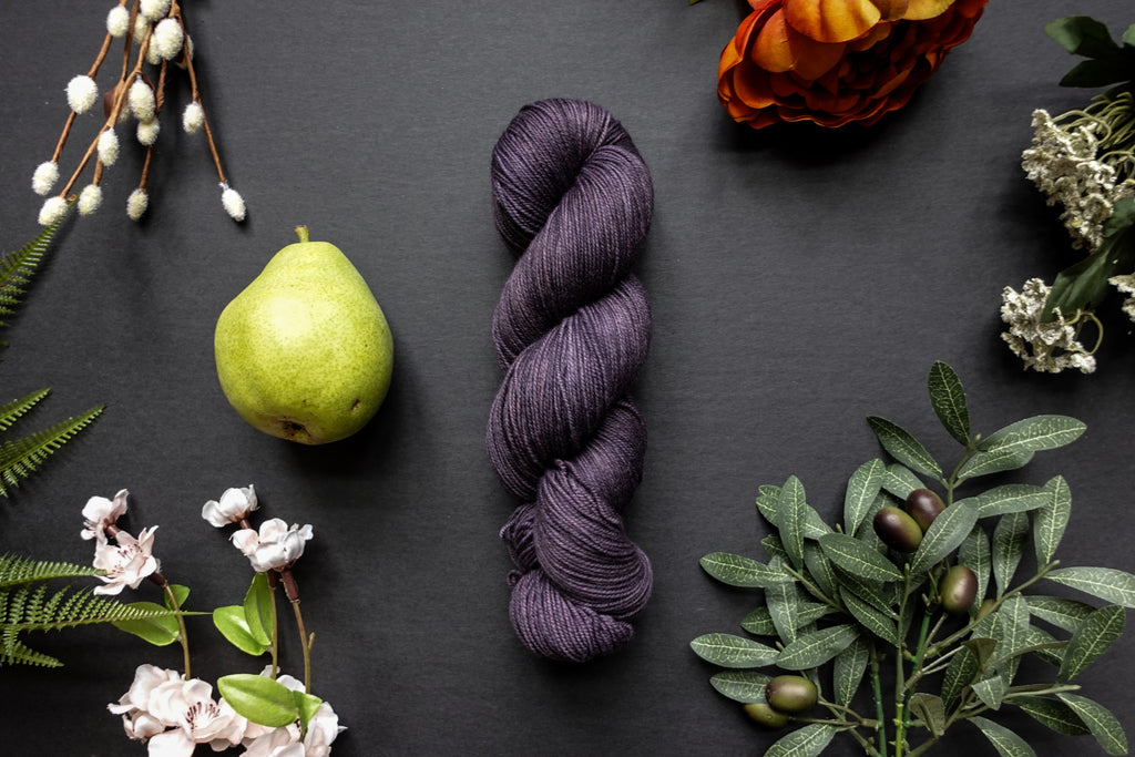 A skein of deep purple sock weight yarn lies on a black surface. It's surrounded by flowers, branches, an orange rose, and a pear.