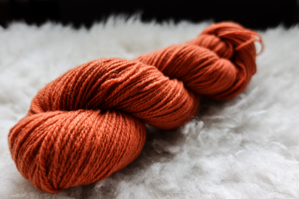 A natural skein of yarn has been hand dyed orange-red. It lays on a sheepskin and is seen from the side.