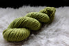 A skein of natural yarn has been hand dyed bright green. It lays on a sheepskin and is pictured from the side.