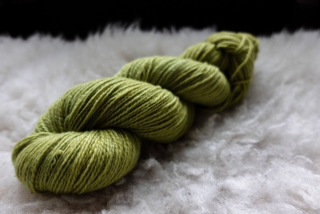 A skein of natural yarn has been hand dyed bright green. It lays on a sheepskin and is pictured from the side.