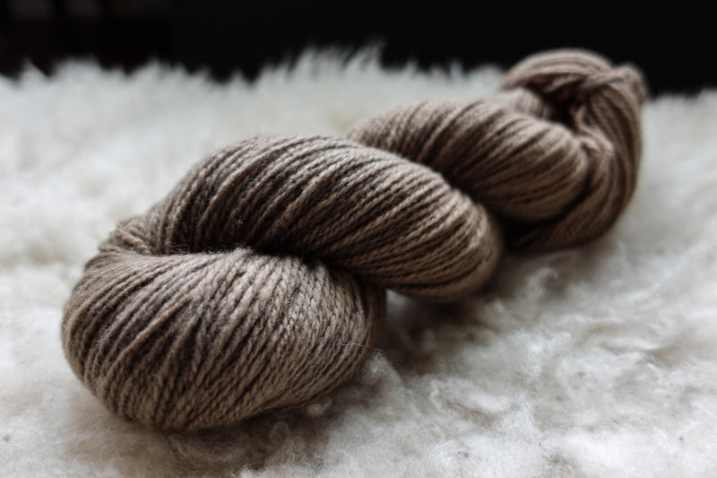 A natural skein of yarn has been hand dyed a light brown. It lays on a sheepskin and is seen from the side.