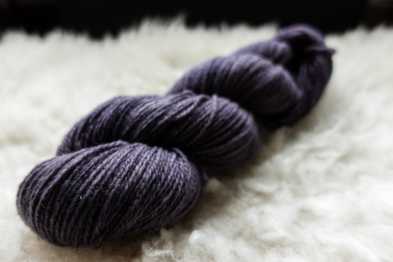 A natural skein of yarn has been hand dyed a deep purple. It lays on a sheepskin and is seen from the side.