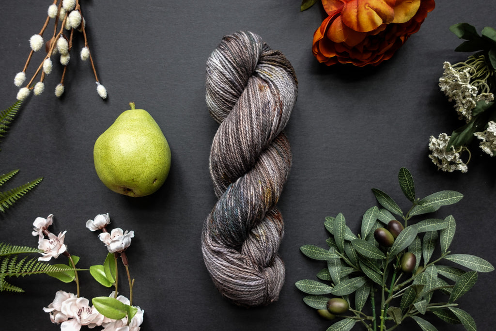 A variegated skein of brown, white, and beige sport weight yarn lies on a black surface. It's surrounded by flowers, branches, an orange rose, and a pear.