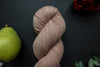 Seen close up, and light pink-beige skein of hand dyed yarn lays on a black surface next to an orange-red flower and a pear.