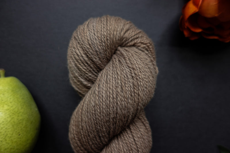 Seen close up, a light brown skein of hand dyed yarn lays on a black surface next to an orange-red flower and a pear.