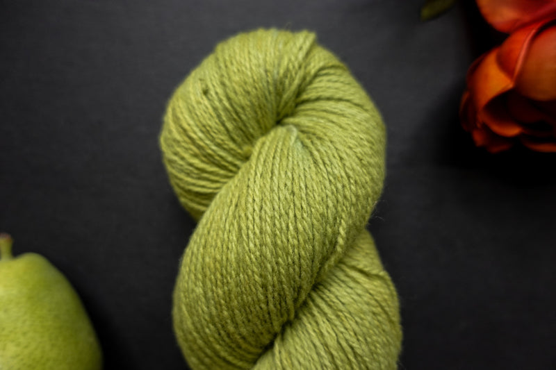 A bright green skein of naturally dyed yarn is seen close up. It lays on a black surface next to an orange-red flower and a pear.