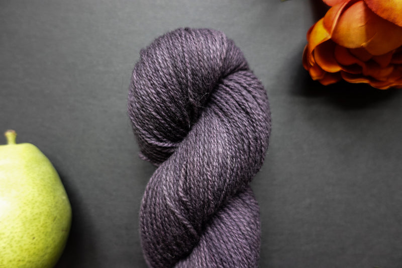 Seen up close, a deep purple skein of hand dyed yarn lays on a black surface next to and orange-red flower and a pear.