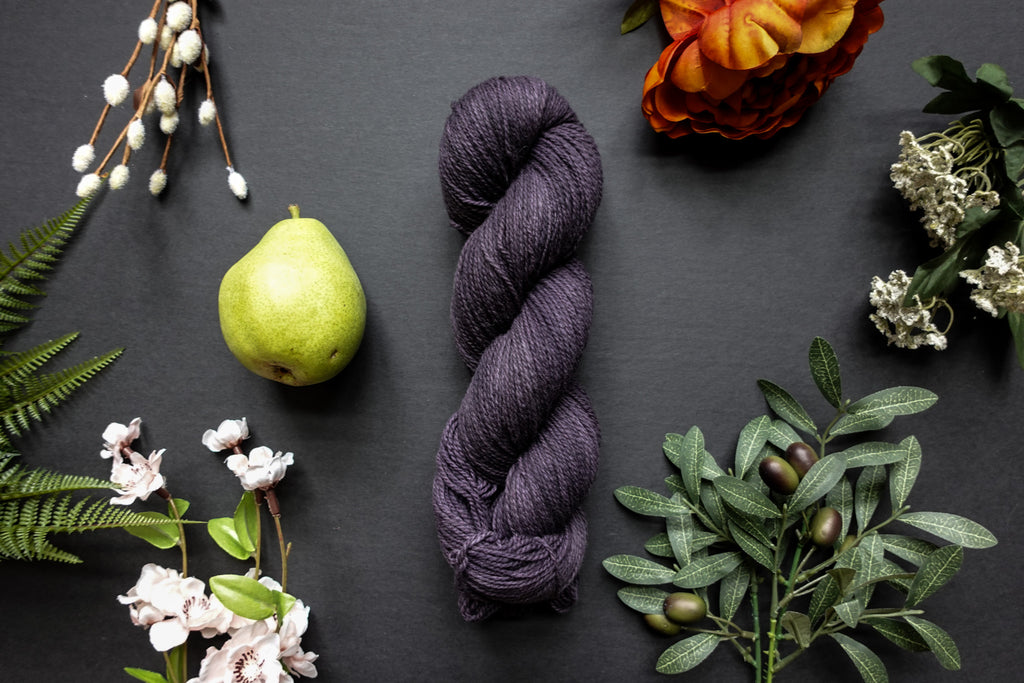 A skein of deep purple sport weight yarn lies on a black surface. It's surrounded by flowers, branches, an orange rose, and a pear.