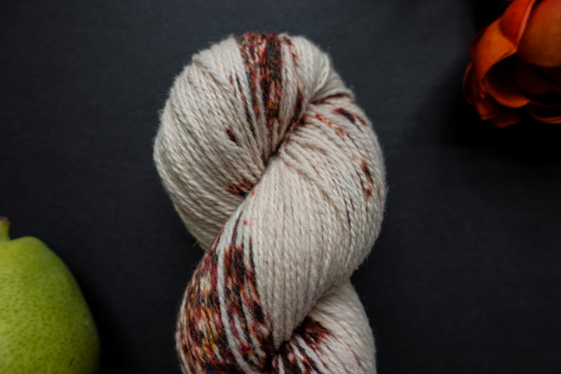 Seen close up, a naturally dyed skein of white yarn with burgundy speckles lays on a black surface next to an orange-red flower and a pear.