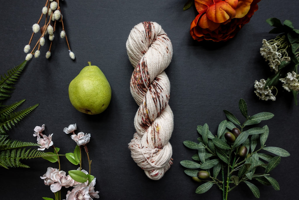 A skein of white sport weight yarn with burgundy speckles lies on a black surface. It's surrounded by flowers, branches, an orange rose, and a pear.