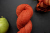 Seen close up, a hand dyed skein of orange-red yarn lays on a black surface next to a matching flower and a pear.