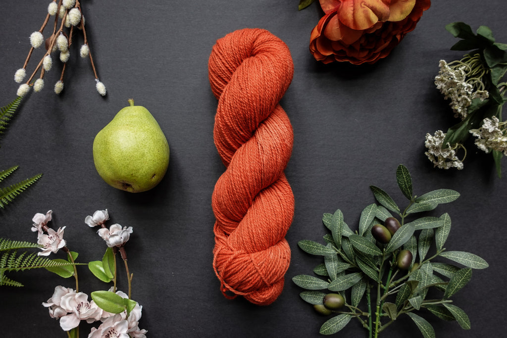 A orange-red skein of sport weight yarn lies on a black surface. It's surrounded by flowers, branches, an orange rose, and a pear.