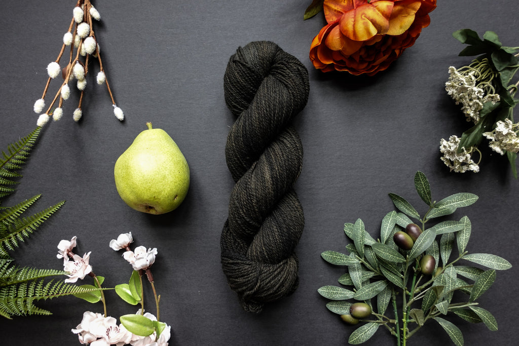 A dark, muddy green, almost black, skein of sport weight yarn lies on a black surface. It's surrounded by flowers, branches, an orange rose, and a pear.