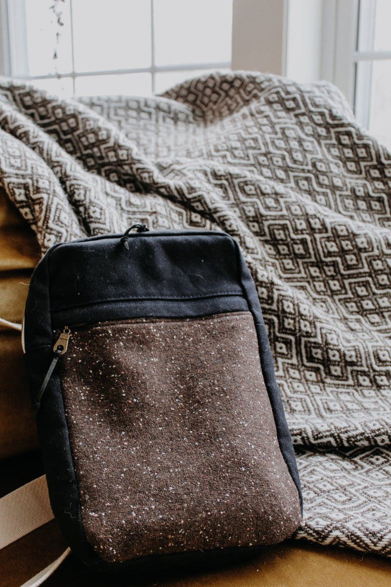 Seen from an angle, a black canvas and brown tweed yarn project bag leans against a blanket. It has a front zippered pockets and a white sling strap.