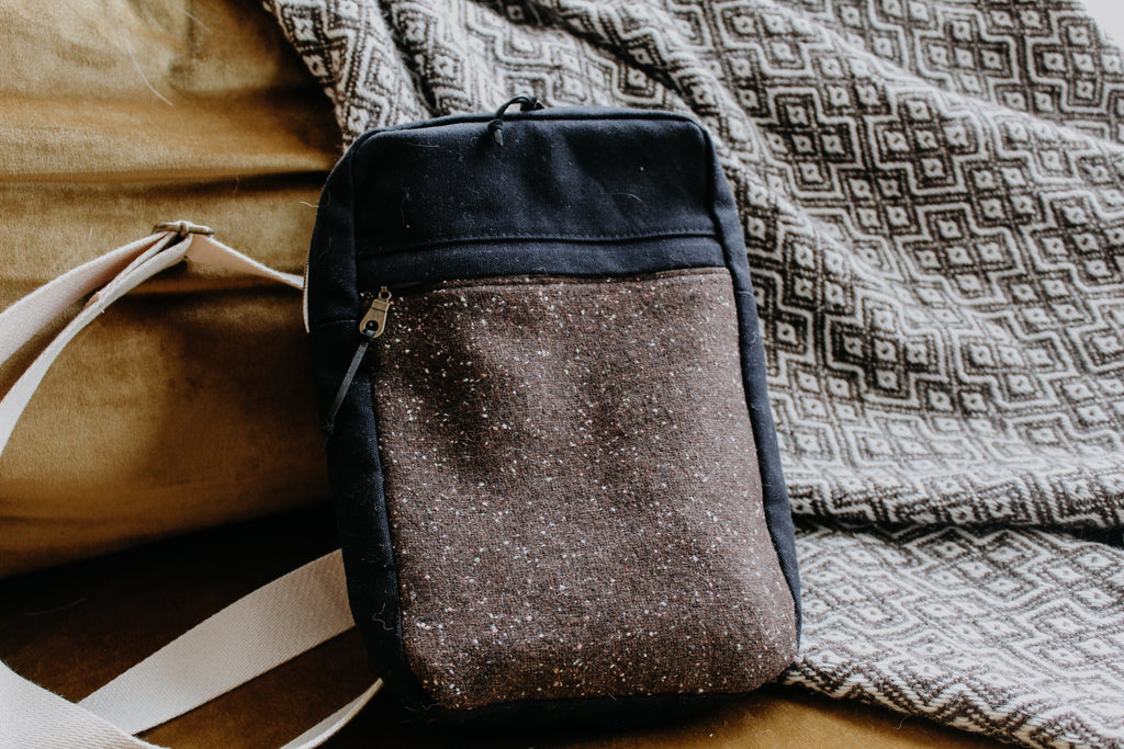 A sling style knitting project bag leans against a blanket. The exterior is made of black canvas and a brown and white handwoven tweed. It has a front zippered pouch and a long white sling strap.