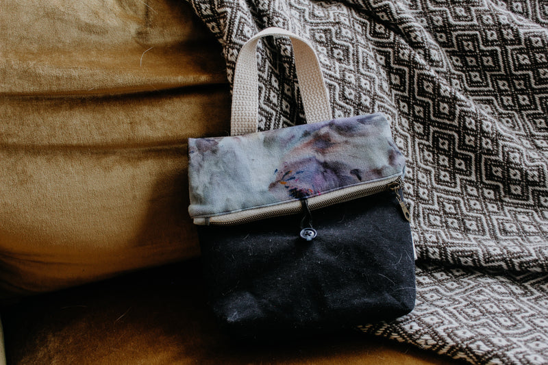 A black and light blue hand dyed project bag leans against a blanket. The top closes with a zipper and button clasp, and has a white handle.