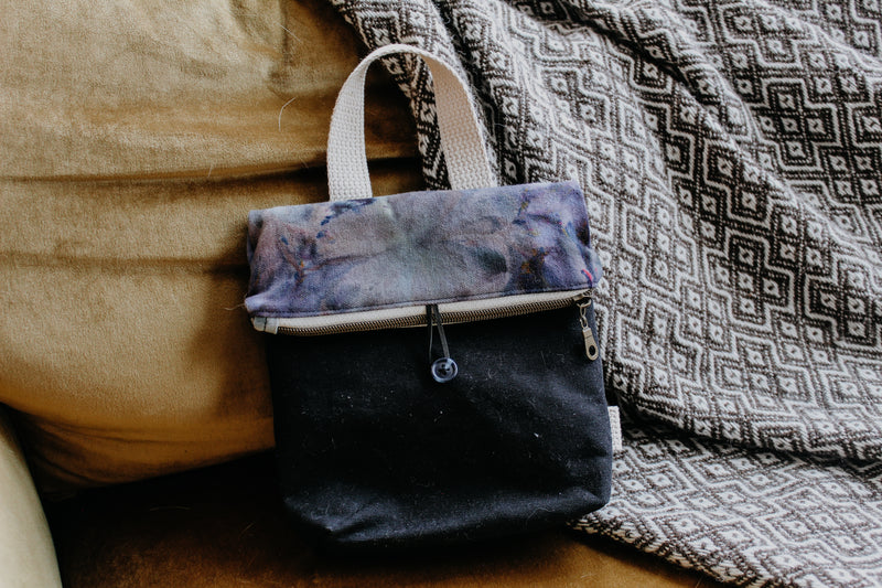 A knitting project bag leans against a blanket. The top is dyed a light blue and purple mix, and the bottom is black canvas. It has a zipper closure, a button clasp, and a white woven handle.