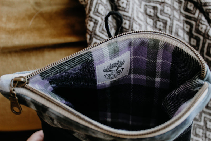 A project bag is open to view the inside. It is lined with a black, white, and purple flannel fabric, and a small version of the Forest Lane Fiber Co. logo has been stitched inside.