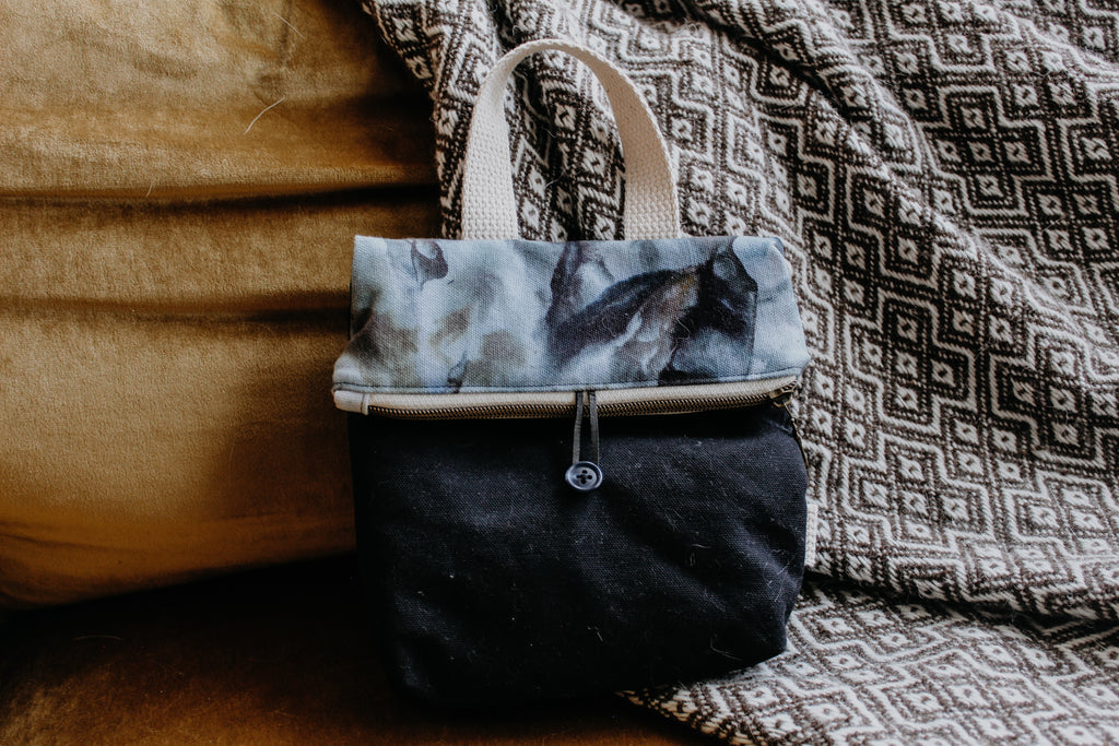 A knitting project bag leans against a blanket. The exterior is ice dyed in a light blue and black mix, contrasting with black canvas. It has a zipper closure and a white handle.